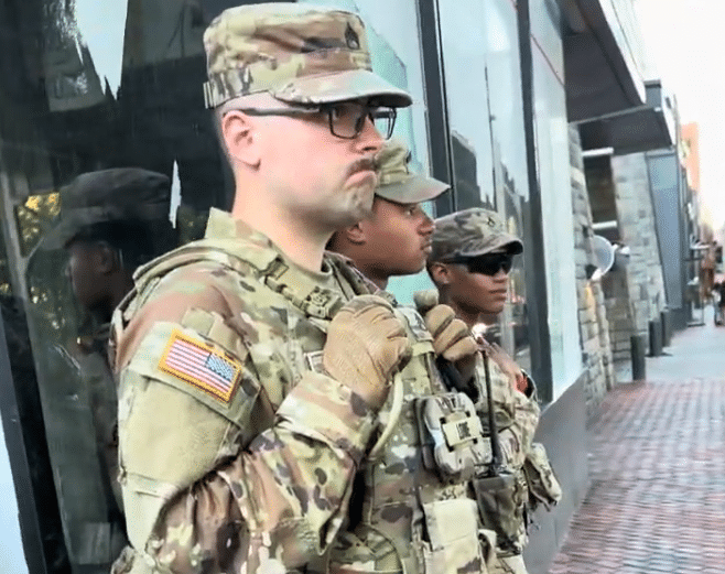 A line of three Michigan National Guard members stands on a brick sidewalk alongside a building's glass storefront. They are wearing matching camouflage utility uniforms and patrol caps, complete with tactical vests, tan combat gloves, and communication gear.