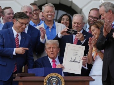 Donald Trump sits at a desk holding up a signed document, surrounded by a smiling crowd of lawmakers clapping and taking photos.