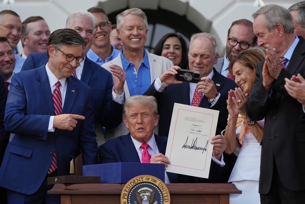 Donald Trump sits at a desk holding up a signed document, surrounded by a smiling crowd of lawmakers clapping and taking photos.