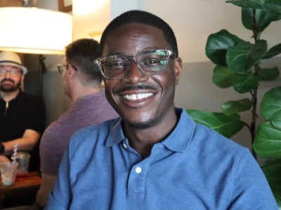 David Crowley smiles broadly while wearing glasses and a blue polo shirt, with other people blurred in the background.