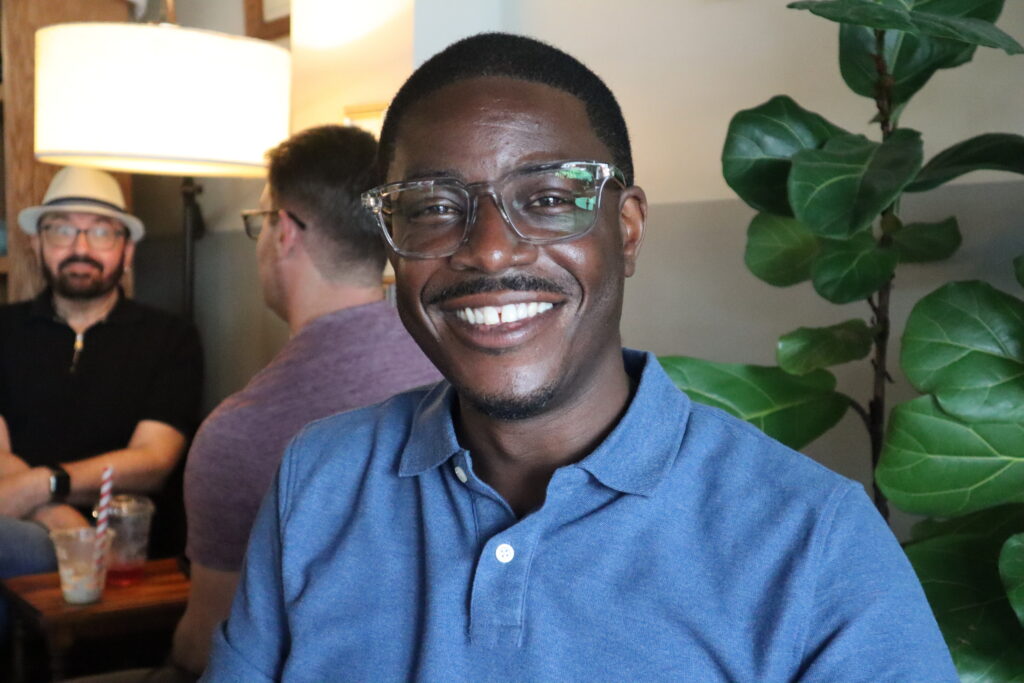 David Crowley smiles broadly while wearing glasses and a blue polo shirt, with other people blurred in the background.