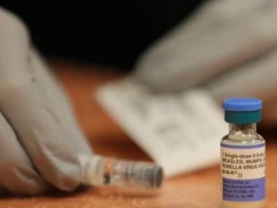 Gloved hands prepare a syringe next to a small vial labeled "MEASLES, MUMPS, AND RUBELLA VIRUS VACCINE".