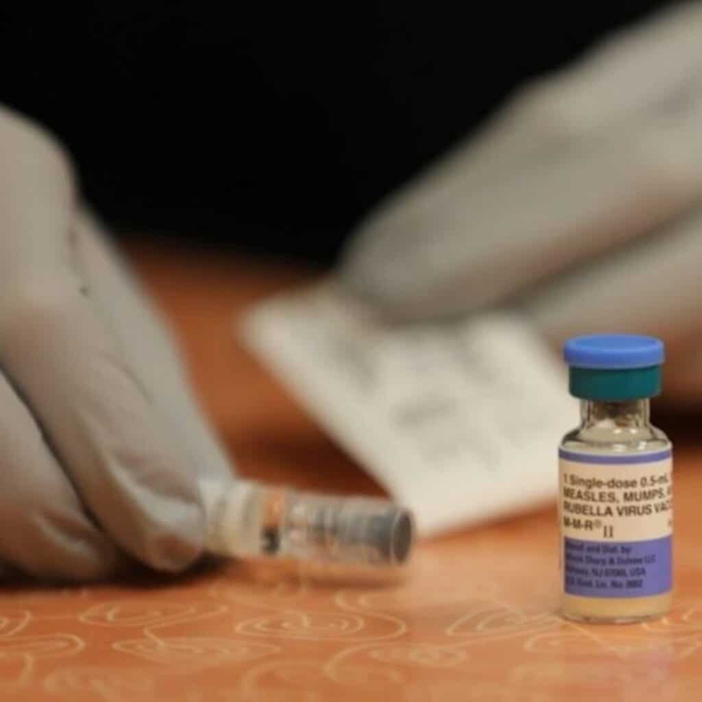 Gloved hands prepare a syringe next to a small vial labeled "MEASLES, MUMPS, AND RUBELLA VIRUS VACCINE".
