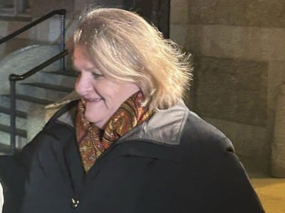 Hannah Dugan walks outdoors in a dark coat and patterned scarf, looking slightly down and smiling.