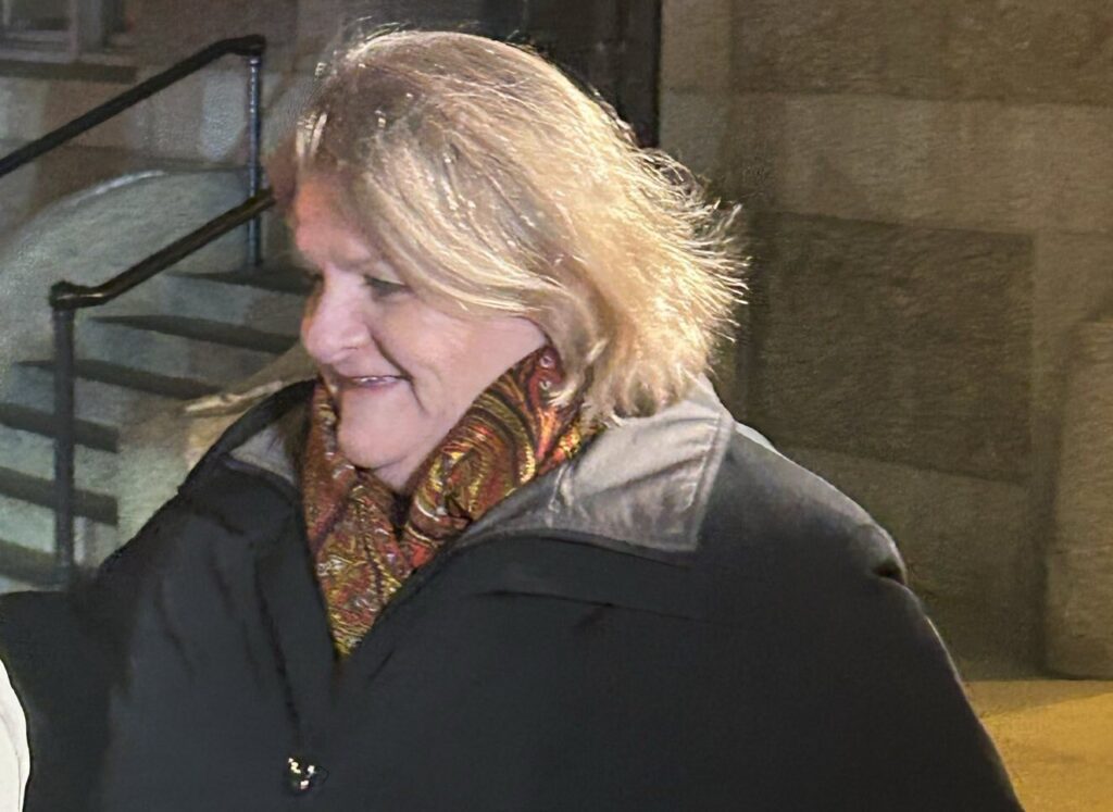 Hannah Dugan walks outdoors in a dark coat and patterned scarf, looking slightly down and smiling.
