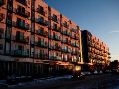 Warm sunset light illuminates the facade and balconies of a large apartment building along a snowy city street.