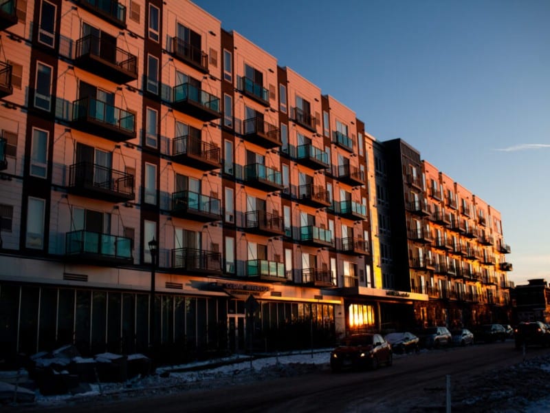 Warm sunset light illuminates the facade and balconies of a large apartment building along a snowy city street.