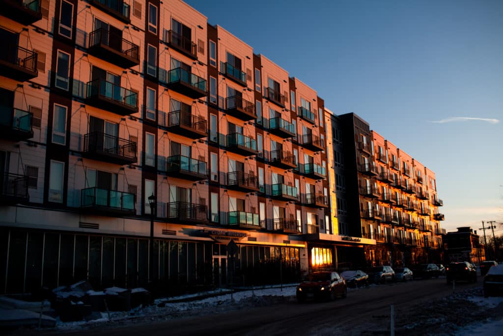 Warm sunset light illuminates the facade and balconies of a large apartment building along a snowy city street.