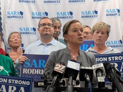 Sara Rodriguez speaks at a press conference microphone stand, surrounded by supporters holding campaign signs.