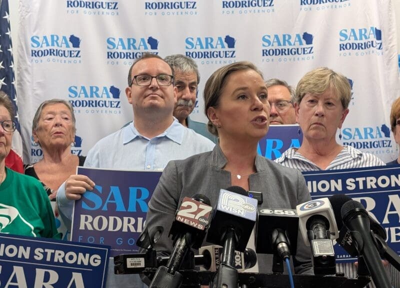 Sara Rodriguez speaks at a press conference microphone stand, surrounded by supporters holding campaign signs.