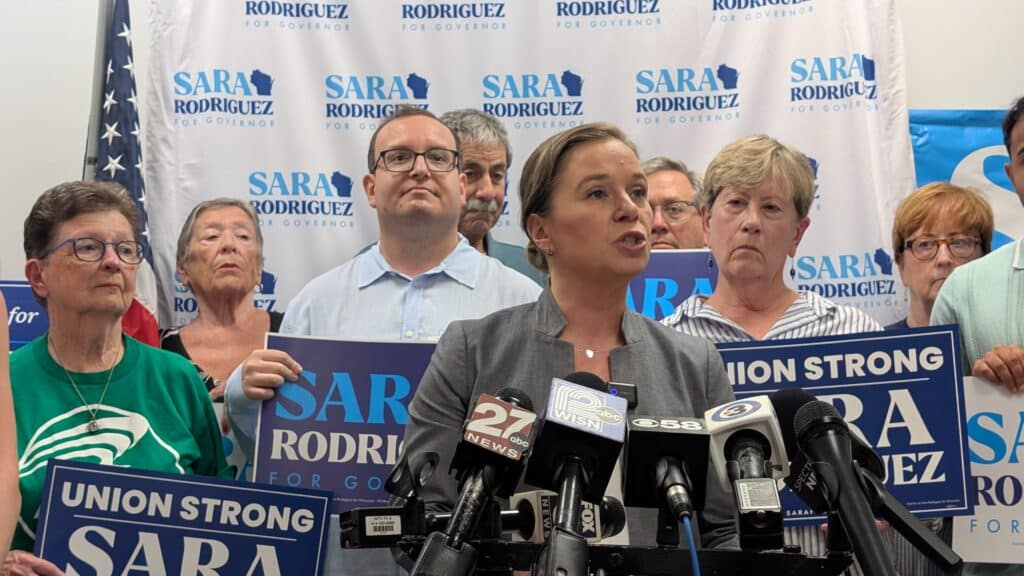 Sara Rodriguez speaks at a press conference microphone stand, surrounded by supporters holding campaign signs.