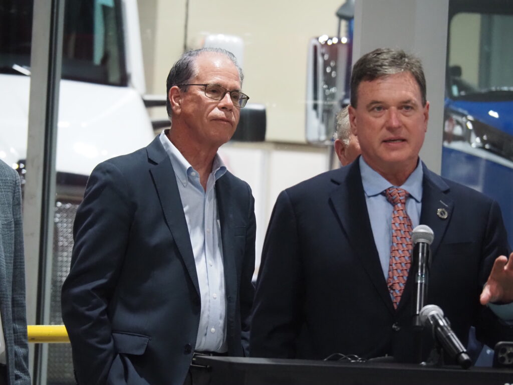 Governor Mike Braun stands behind Attorney General Todd Rokita as Rokita speaks at a microphone stand inside a truck dealership.
