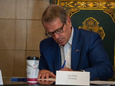 Governor Jim Pillen sits at a table and uses a blue pen to sign paperwork behind a nameplate reading "Jim Pillen Governor."
