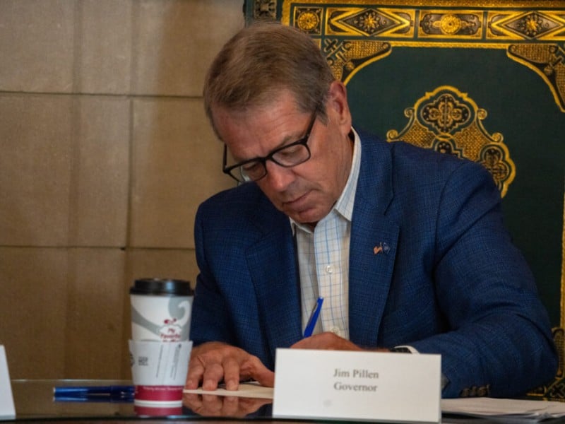 Governor Jim Pillen sits at a table and uses a blue pen to sign paperwork behind a nameplate reading "Jim Pillen Governor."