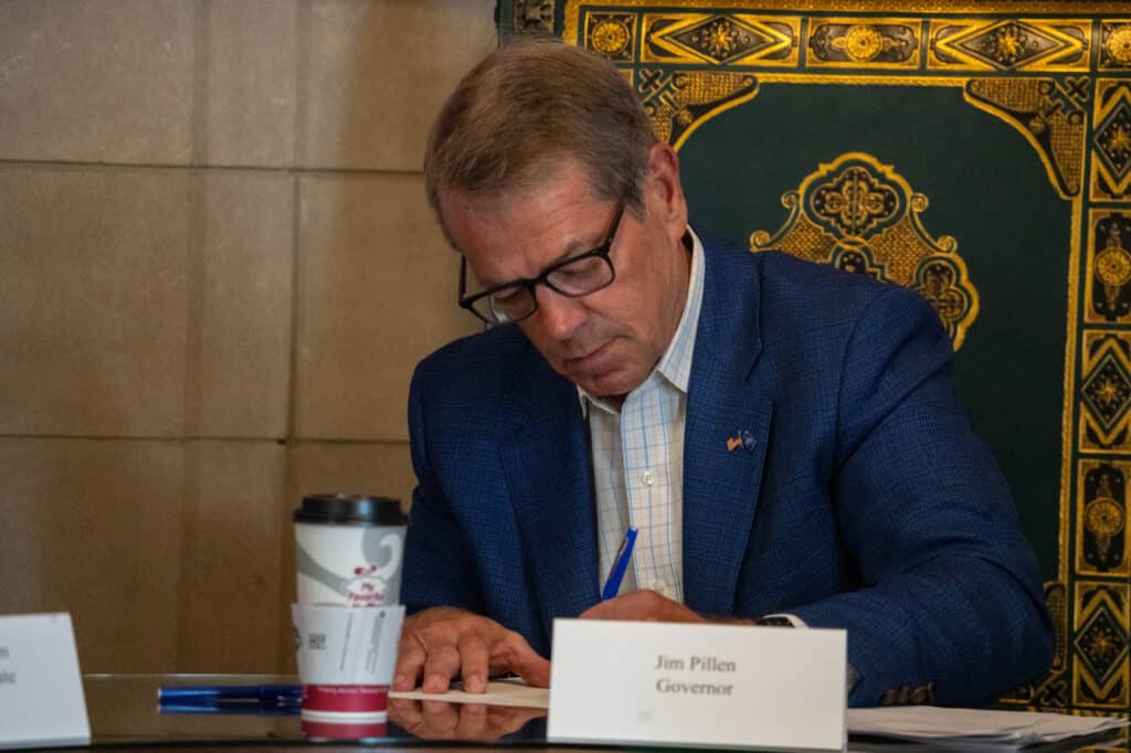 Governor Jim Pillen sits at a table and uses a blue pen to sign paperwork behind a nameplate reading "Jim Pillen Governor."