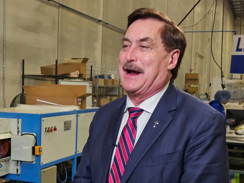 Mike Lindell speaks inside a manufacturing facility with machinery in the background.