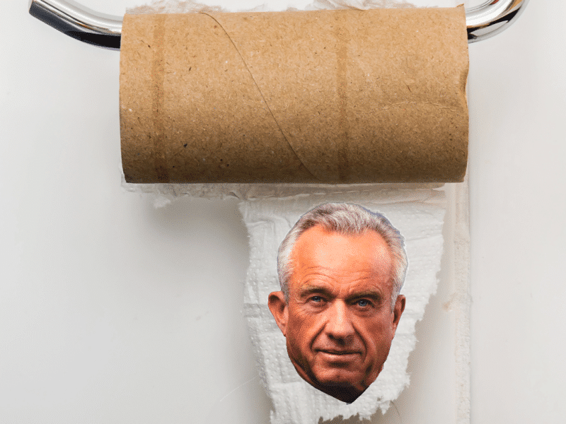 An empty, brown cardboard toilet paper roll on a chrome holder with a small piece of paper featuring Robert F. Kennedy Jr.'s face.