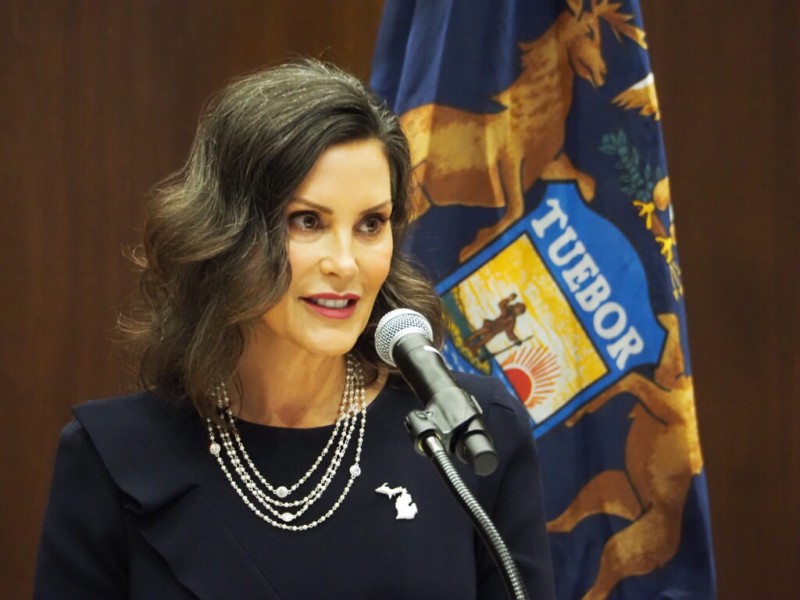Michigan Governor Gretchen Whitmer speaks at a microphone, with the Michigan state flag in the background