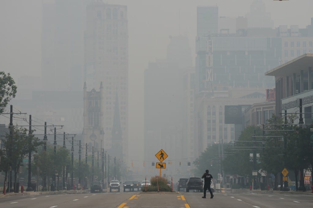 Thick smoke and haze shroud Detroit skyscrapers and a wide street where a pedestrian walks.