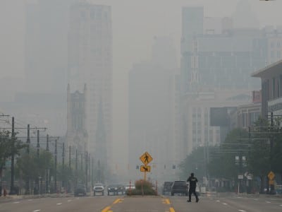 Thick smoke and haze shroud Detroit skyscrapers and a wide street where a pedestrian walks.