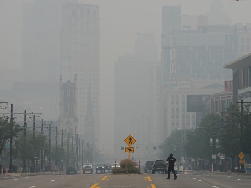 Thick smoke and haze shroud Detroit skyscrapers and a wide street where a pedestrian walks.