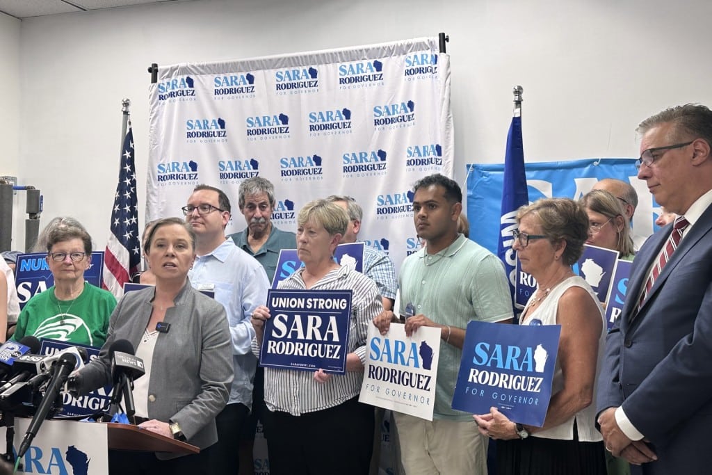 Sara Rodriguez speaks at a podium, surrounded by supporters holding "Sara Rodriguez" campaign signs.