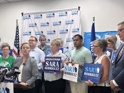Sara Rodriguez speaks at a podium, surrounded by supporters holding "Sara Rodriguez" campaign signs.