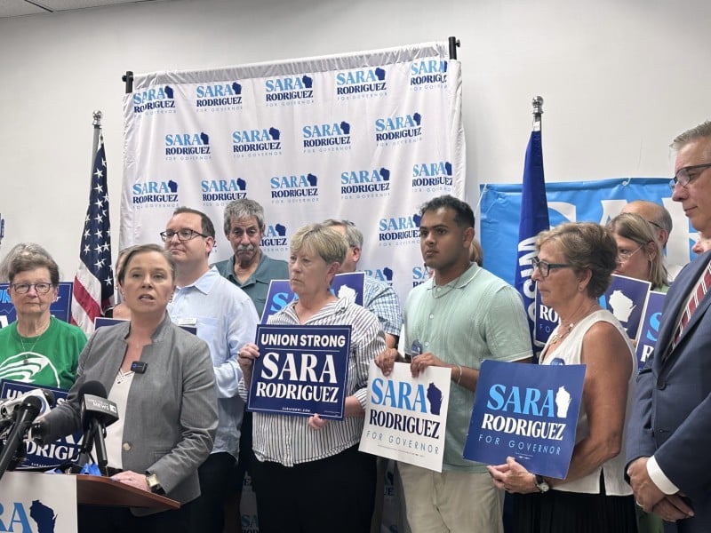 Sara Rodriguez speaks at a podium, surrounded by supporters holding "Sara Rodriguez" campaign signs.