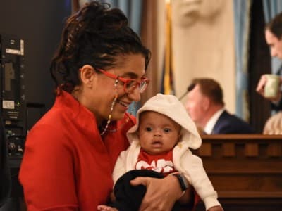 Dr. Mona Hanna smiles warmly while holding a baby wearing a white hooded jacket.