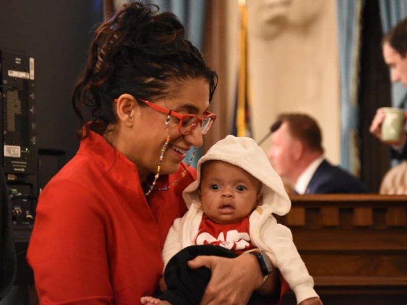 Dr. Mona Hanna smiles warmly while holding a baby wearing a white hooded jacket.