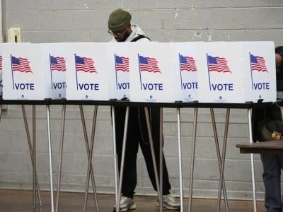 A man wearing a beanie stands behind a row of white voting privacy booths labeled "VOTE" with American flags while filling out a ballot.