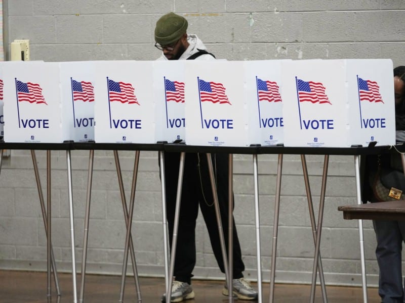 A man wearing a beanie stands behind a row of white voting privacy booths labeled "VOTE" with American flags while filling out a ballot.