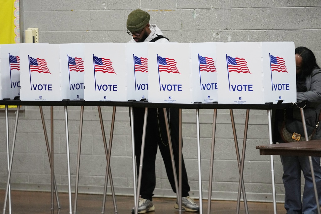 A man wearing a beanie stands behind a row of white voting privacy booths labeled "VOTE" with American flags while filling out a ballot.