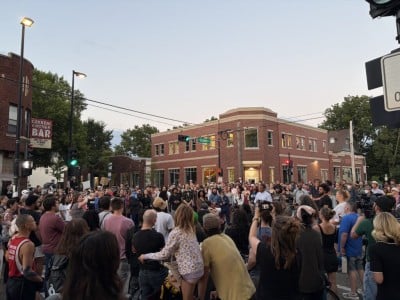 A large crowd of people gathers in a street intersection at dusk outside brick buildings.