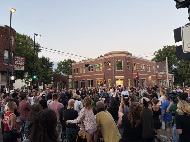A large crowd of people gathers in a street intersection at dusk outside brick buildings.
