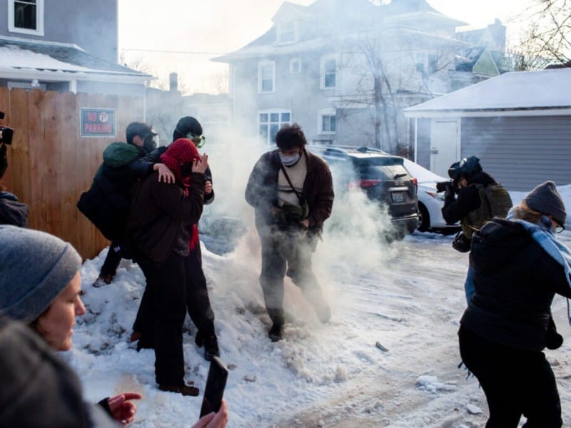 Demonstrators in a snowy alley cover their faces and scatter through thick tear gas smoke