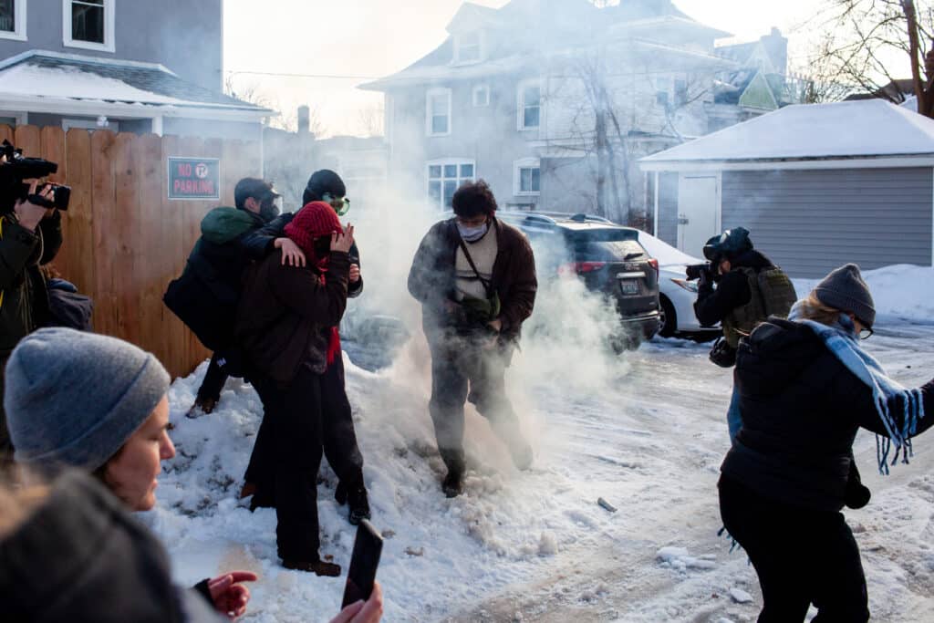 Demonstrators in a snowy alley cover their faces and scatter through thick tear gas smoke