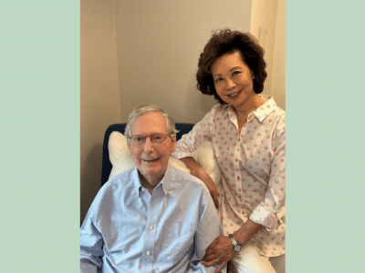 U.S. Sen. Mitch McConnell sits smiling next to his wife, Elaine Chao, who stands beside him with a hand on his arm.