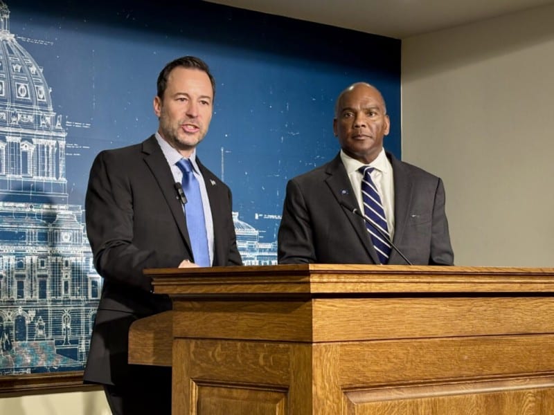 Brian Nicholson speak at a podium while Kendall Qualls stands beside him in front of a Capitol backdrop