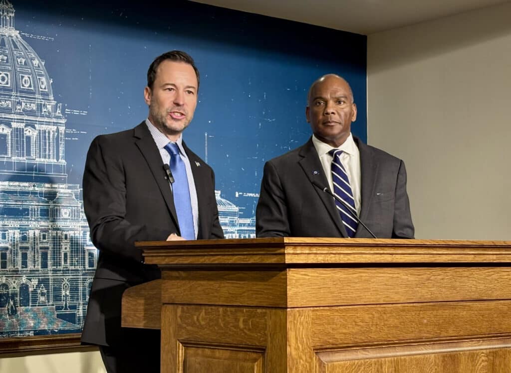 Brian Nicholson speak at a podium while Kendall Qualls stands beside him in front of a Capitol backdrop