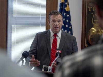 Kansas Attorney General Kris Kobach gestures while speaking into microphones at a press conference.
