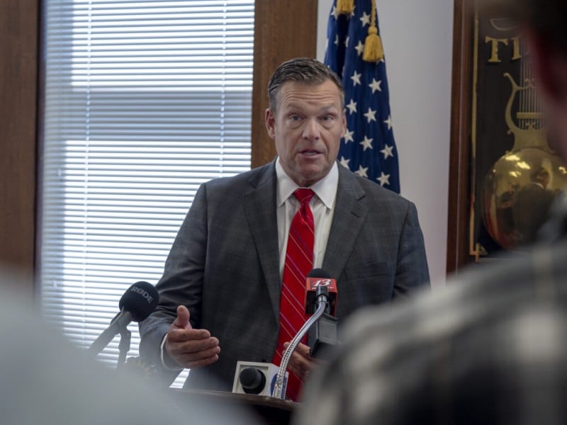 Kansas Attorney General Kris Kobach gestures while speaking into microphones at a press conference.