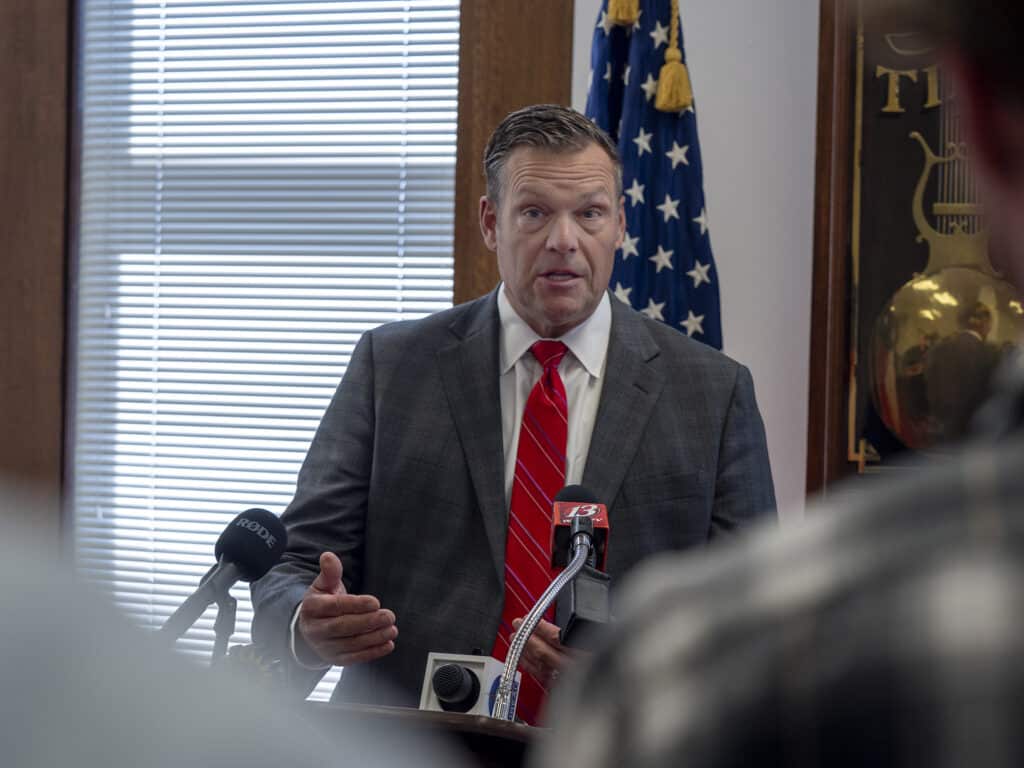 Kansas Attorney General Kris Kobach gestures while speaking into microphones at a press conference.