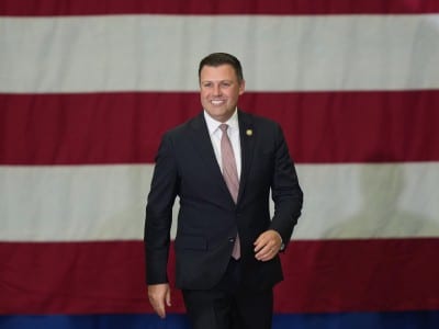 Rep. Mackenzie, smiling and wearing a blue suit, walks forward in front of a large American flag backdrop.