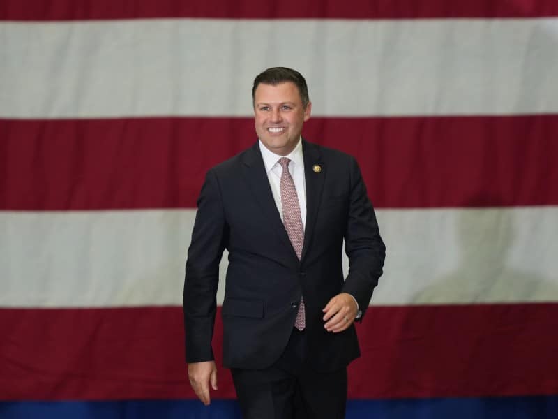 Rep. Mackenzie, smiling and wearing a blue suit, walks forward in front of a large American flag backdrop.
