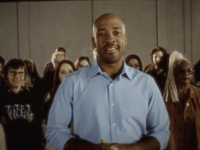 Mandela Barnes smiles directly at the camera, standing in front of a group of supporters indoors.