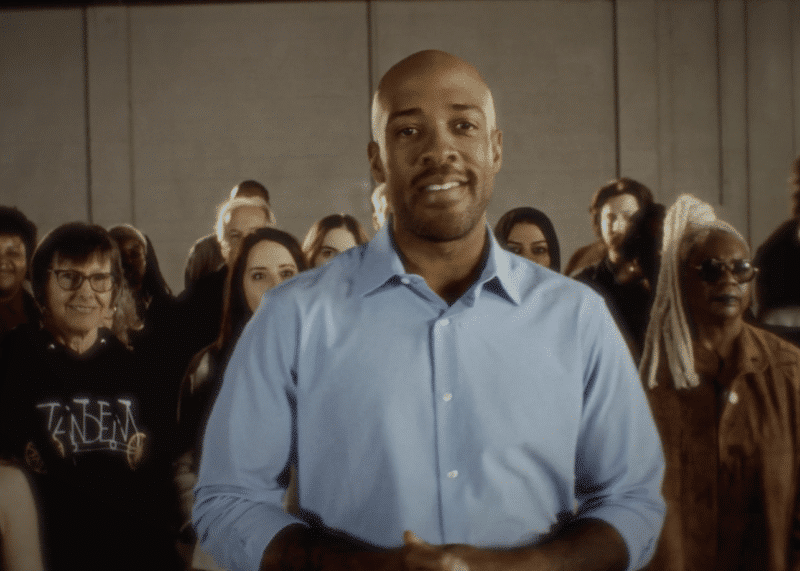 Mandela Barnes smiles directly at the camera, standing in front of a group of supporters indoors.