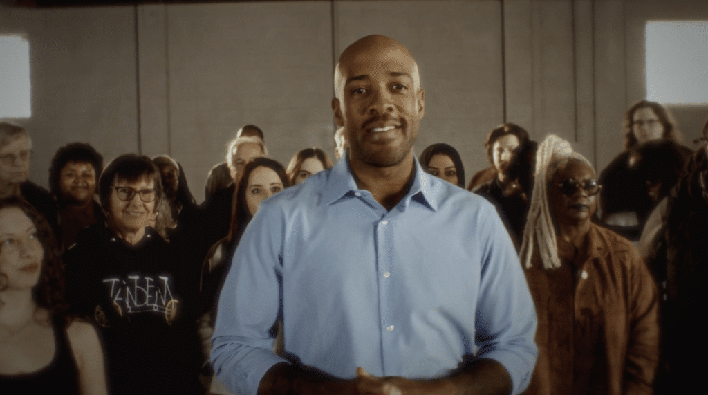 Mandela Barnes smiles directly at the camera, standing in front of a group of supporters indoors.