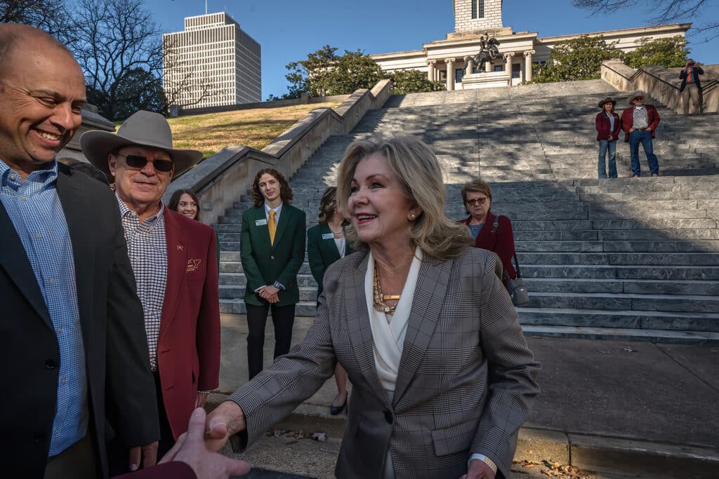 U.S. Sen. Marsha Blackburn smiles and shakes hands on the steps of a grand stone building with a crowd behind her.