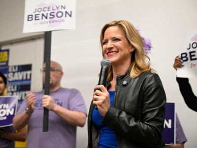 Jocelyn Benson smiles while speaking into a microphone, surrounded by supporters holding "Jocelyn Benson for Governor" signs.